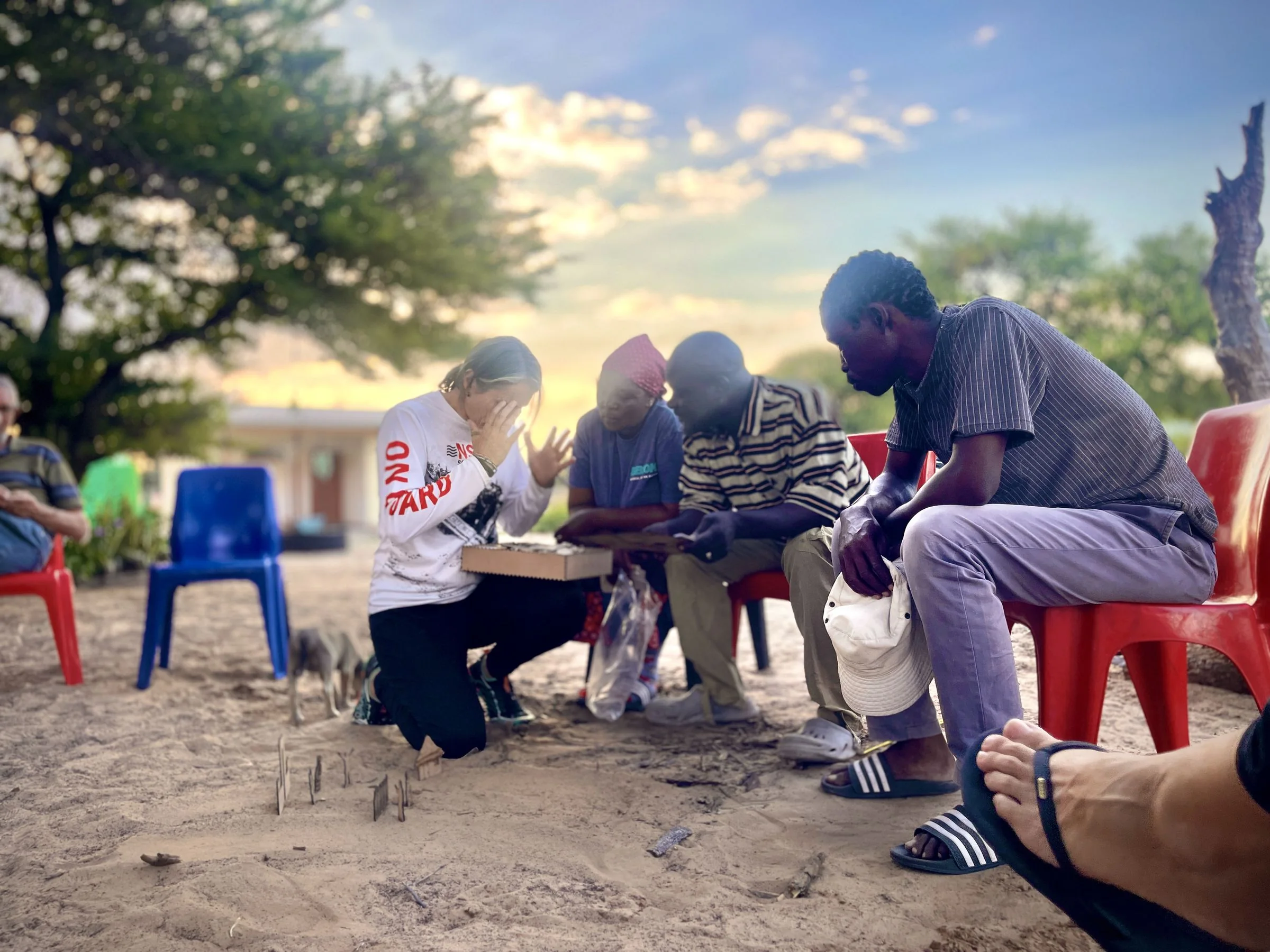 Sarah kneels on the sand with her hands raised to her face beside a box of small wooden figures, while three people sit close around her on plastic chairs and look at the figures; more flat wooden figures stand in the sand in front of them, with a tree and a sunset sky behind.